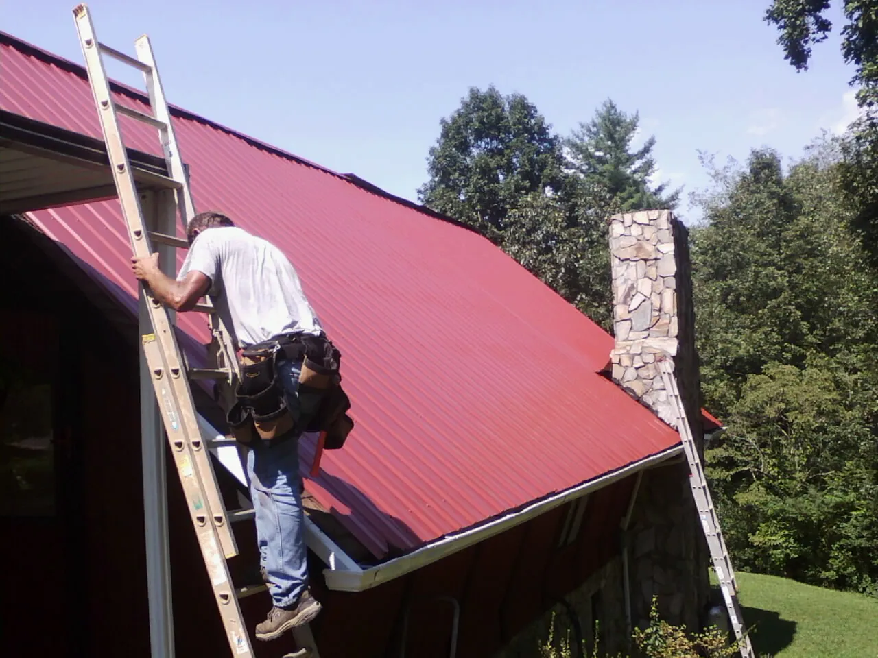 Roofer on a ladder beside a red metal roof and stone chimney — Soffit & Fascia Repair by Roofing Brothers in Culleoka